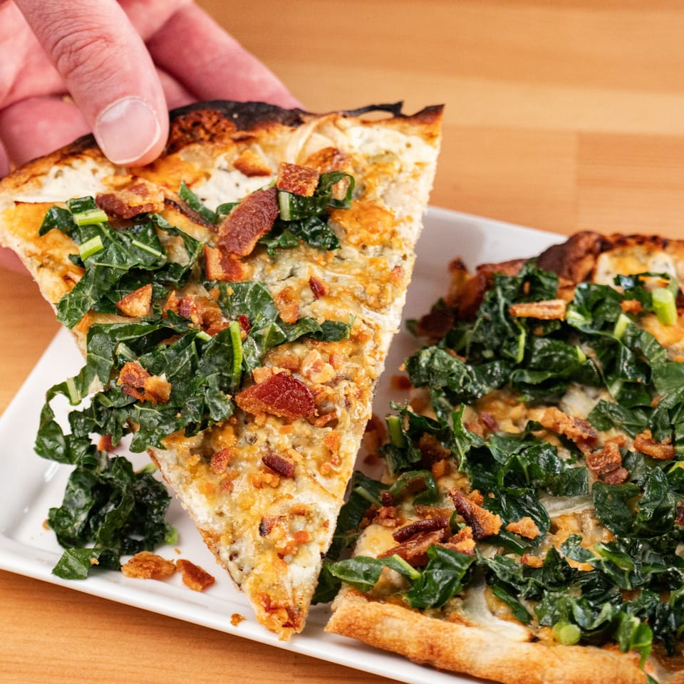 Hand lifting a slice of Milque Toast breakfast pizza with kale, bacon, and cheese.