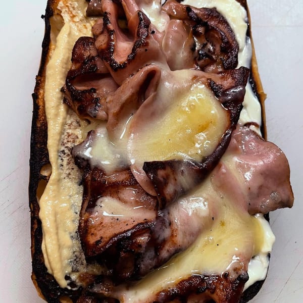 Close-up of a Milque Toast breakfast sandwich with melted cheese, meat, and sauce on toasted bread.