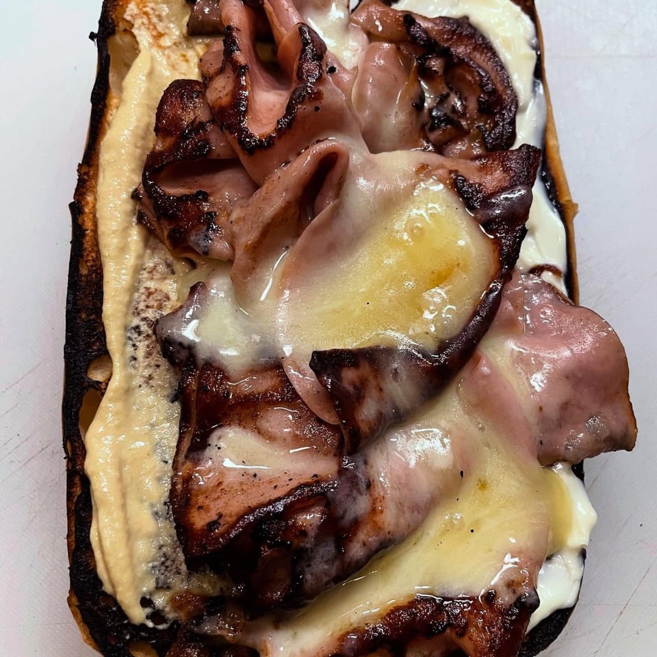 Close-up of a Milque Toast breakfast sandwich with melted cheese, meat, and sauce on toasted bread.