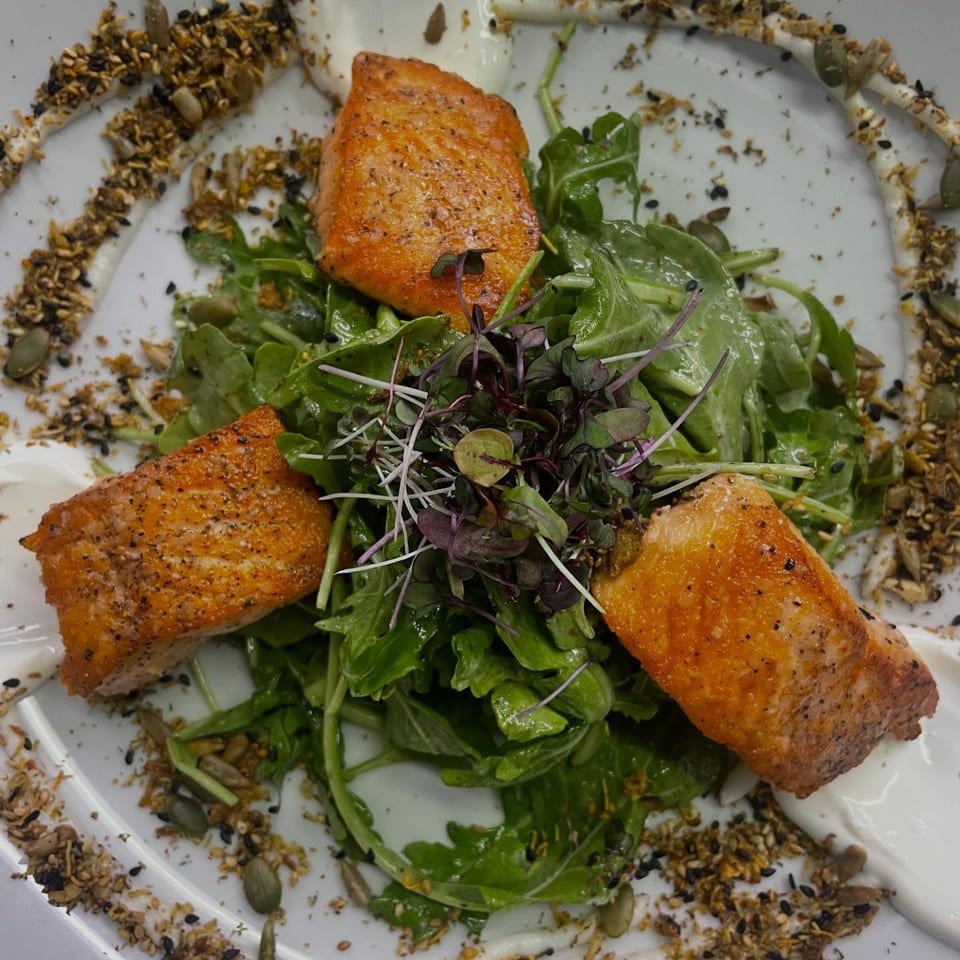 Three pieces of seared salmon on a bed of arugula, garnished with microgreens and seeds.
